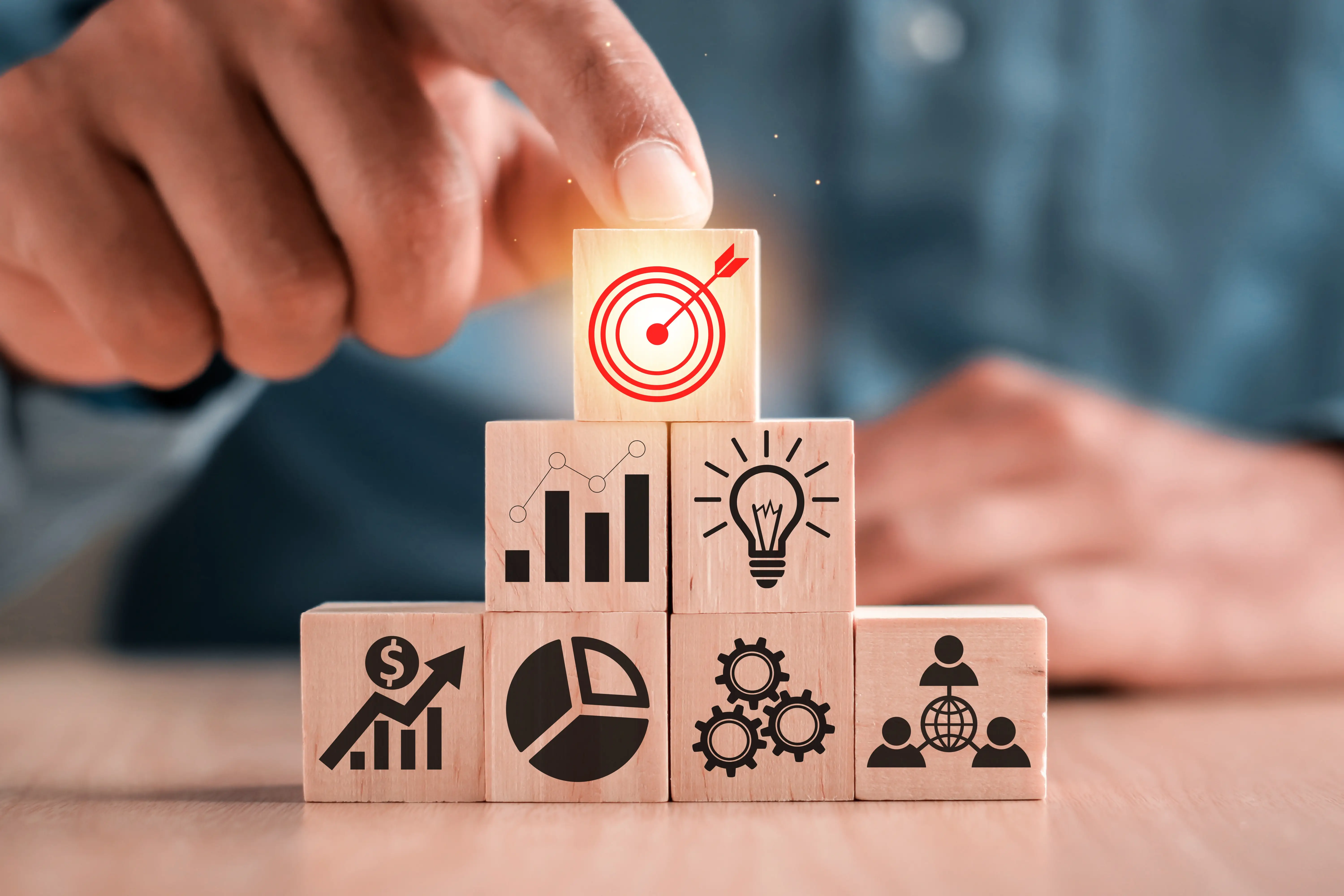 A hand placing a wooden block with a target icon on top of other blocks with business-related symbols, representing strategic goal-setting and achieving results.